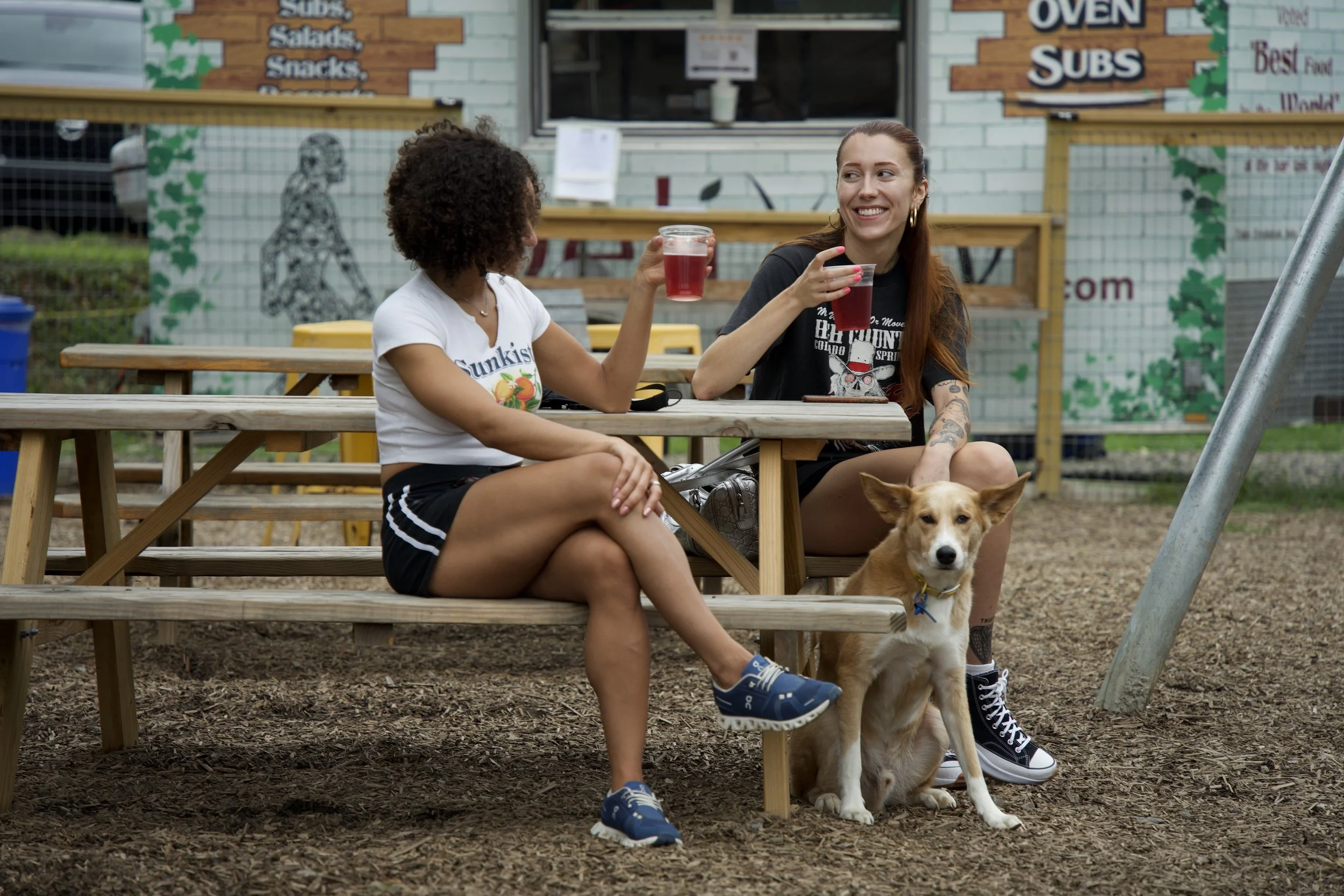 Two women toast drinks at a picnic table while a tan dog sits beside them on wood chips