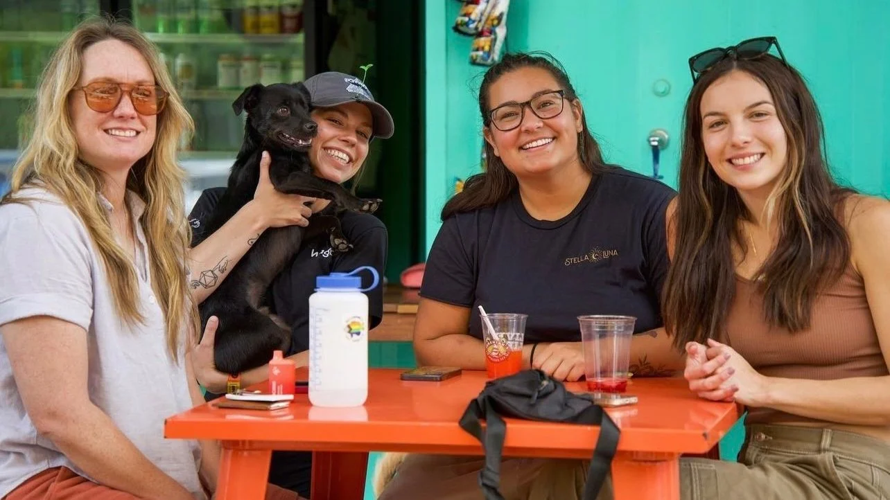 Four smiling women sit at an orange picnic table with a black dog held by one of them, beside cups and a water bottle